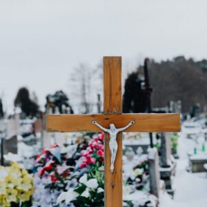 Wintry cemetery scene with a wooden cross in focus and colorful grave flowers.