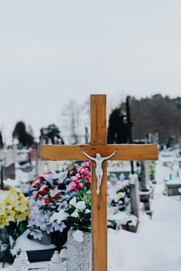 Wintry cemetery scene with a wooden cross in focus and colorful grave flowers.