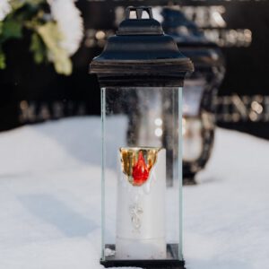 A glass lantern candle on a snow-covered tombstone symbolizes remembrance.